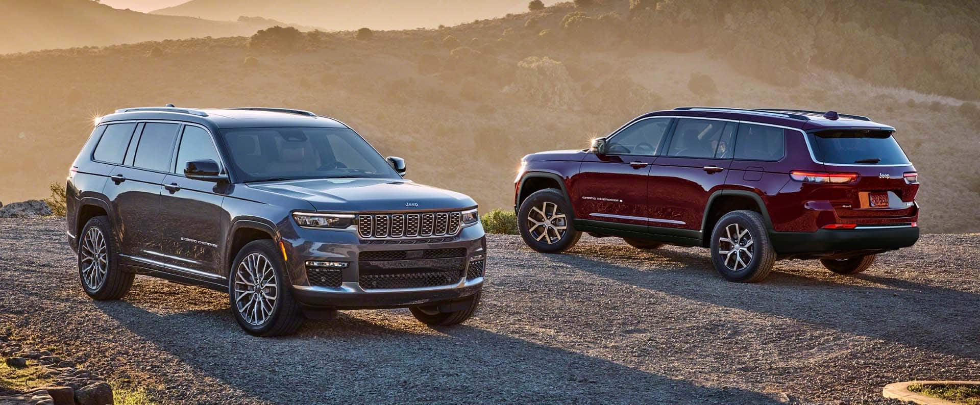 Un Jeep Grand Cherokee Summit Reserve gris y un Jeep Grand Cherokee Limited 2024 rojo estacionado uno frente al otro en una carretera de montaña de ripio al atardecer.