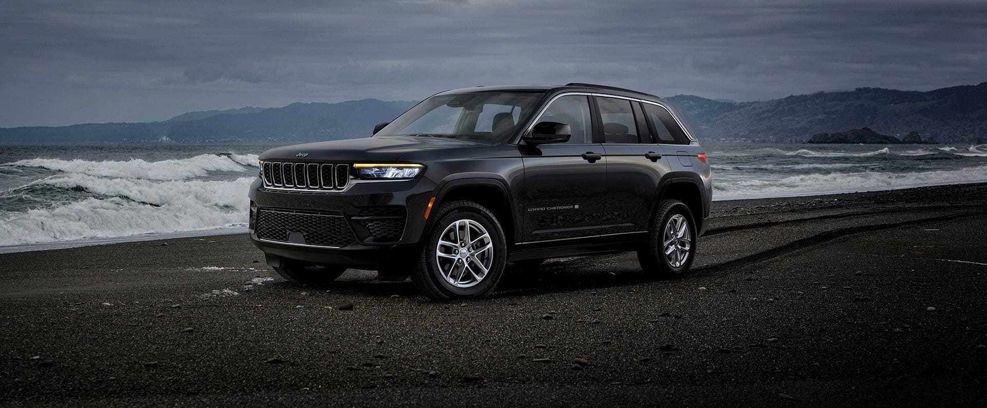 Ángulo delantero del lado del conductor de un Jeep Grand Cherokee Laredo X 2024 negro estacionado en la playa, con un cielo oscuro y tormenta a la distancia.