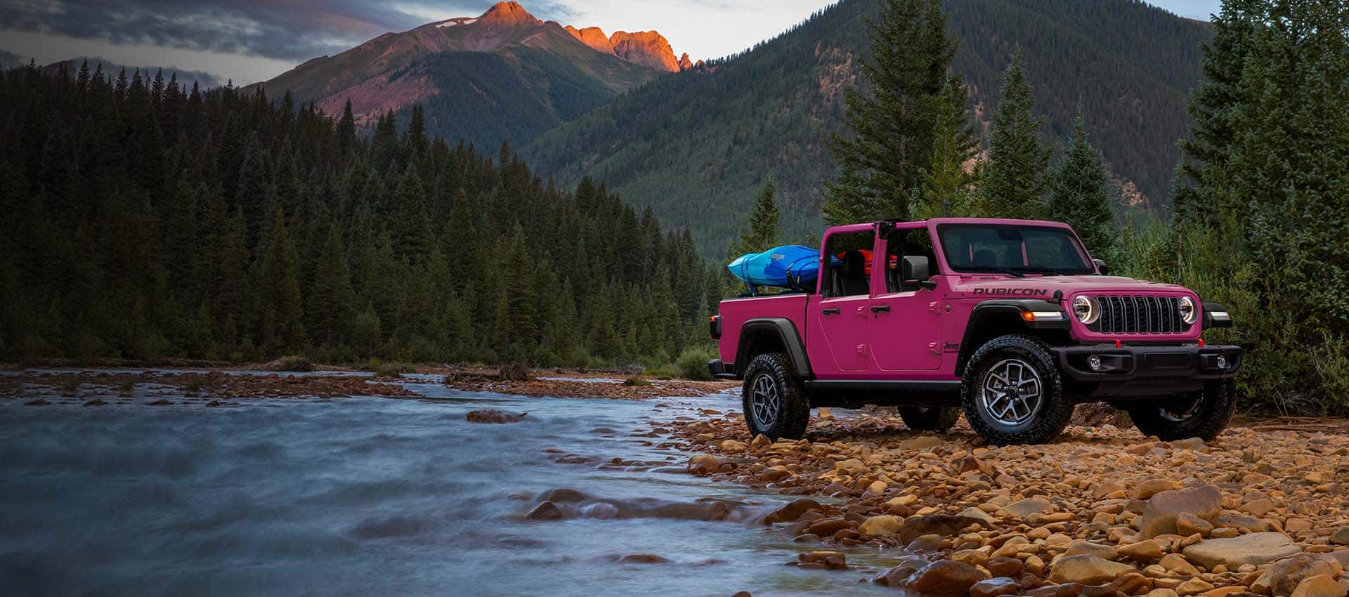 A passenger-side front angle of a pink 2024 Jeep Gladiator Rubicon parked on a bed of rocks beside a stream, with gear in its pickup box and a dense mountain forest in the background.