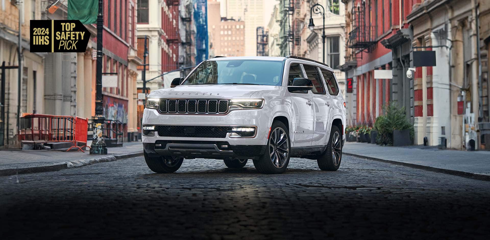 A silver 2024 Jeep Wagoneer Series III parked on a cobblestone street in a city's business district. 2024 IIHS Top Safety Pick.